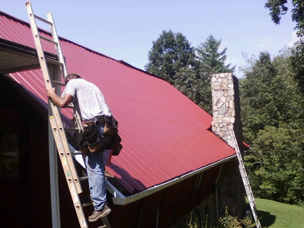Roofer on a ladder beside a red metal roof and stone chimney — Soffit & Fascia Repair by Roofing Brothers in Decherd